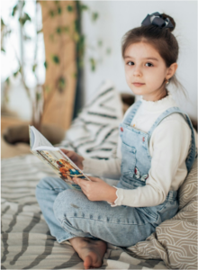 Girl sitting on bed reading a book and looking up at the camera.